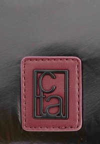 Cuero negro brillante con una etiqueta rectangular burdeos que presenta un logotipo negro en relieve. Bordes cosidos, textura suave y líneas limpias.