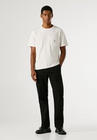 Valge puuvillane T-särk rinnataskuga, millel on sinine tekst, koos mustade sirgete pükste ja mustade nööbiga jalanõudega.