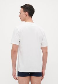 Hvit bomulls t-skjorte med korte ermer, rund hals og avslappet passform. Båret med marineblå shorts, sett bakfra. Glatt tekstur.