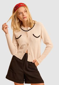 Cardigan beige con rifiniture nere, lavorazione a maglia strutturata e tasche decorative. Abbinato a pantaloni corti neri. Modella tocca i capelli e si trova contro uno sfondo grigio.