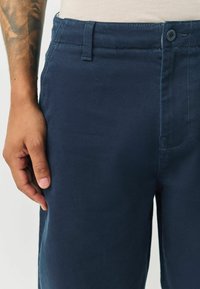 Navy blue cotton pants with a smooth texture, featuring a button closure, belt loops, and visible seam details. Hand resting on the waistband.