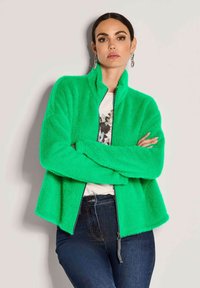 Frau mit dunklem Haar, die silberne Ohrringe trägt, eine leuchtend grüne flauschige Zip-Jacke, ein weißes Hemd und dunkle blaue Jeans anhat, steht mit verschränkten Armen.