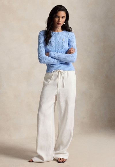 Vrouw met lang donker haar, gekleed in een lichtblauwe kabelgebreide trui en losse witte broek, staand met gekruiste armen tegen een neutrale achtergrond.