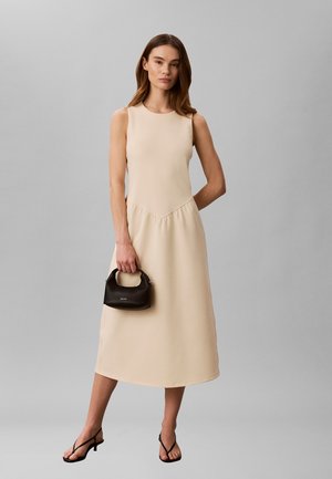 Donna che indossa un vestito midi beige senza maniche, che tiene in mano una piccola borsa nera e indossa sandali neri con cinturini e tacco basso, in piedi davanti a uno sfondo grigio.