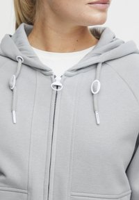 Gray zip-up hoodie with a textured finish, featuring adjustable drawstrings, front pockets, and a regular fit design.