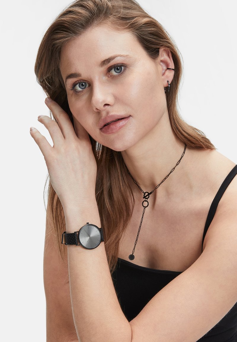 Ronde zwarte klok met een gestructureerde leren band, zilveren wijzerplaat, minimalistisch ontwerp en geen zichtbare cijfers. Het model draagt een zwarte ketting.