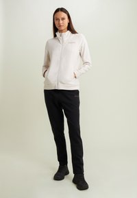 Una donna sta in piedi con le mani in tasca, indossando una giacca crema con zip e pantaloni neri, entrambi con il logo "FREDDY", e delle sneakers nere.