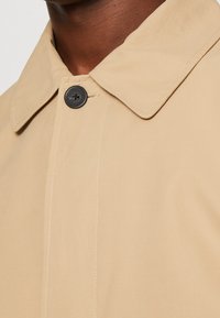 Colletto e bottone superiore di un cappotto beige indossato da una persona di pelle scura, che mette in risalto la texture del tessuto e i dettagli delle cuciture.