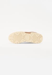 Chaussure de sport avec une tige en suède marron, des accents blancs et une semelle en caoutchouc beige texturée avec un motif de bande de roulement.