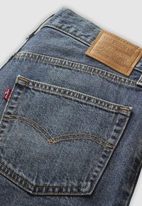 Jean en denim bleu Levi's avec un patch en cuir beige, des coutures visibles et un design classique de poche arrière avec un détail de couture en arc.