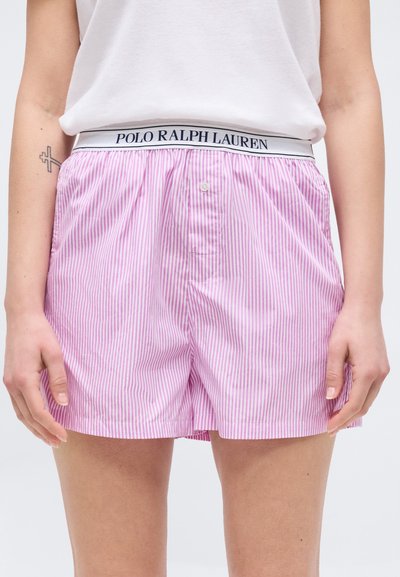 Γυναίκα που φοράει ροζ και λευκά ριγέ σορτς με λευκή μέση που γράφει "Polo Ralph Lauren", συνδυασμένα με απλό λευκό πουκάμισο.