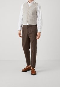 Light beige vest with five buttons over a white shirt, paired with brown trousers and brown slip-on shoes featuring cut-out details.