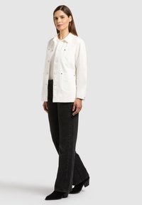Femme debout, portant une veste blanche à boutons, un haut blanc, un jean noir à jambes larges et des bottines à talons noires sur un fond gris clair.