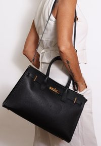 Vrouw in een witte outfit die een grote zwarte leren handtas met gouden sluiting en bovenhandvatten over haar schouder draagt.