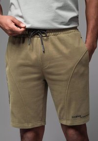 Shorts vert olive en tissu doux avec une taille élastique et un cordon de serrage ; dotés de poches latérales et d'un détail logo subtil sur la jambe.