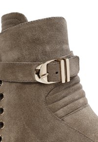 Botte de cheville en daim taupe avec une accent en boucle dorée, un revêtement texturé et des œillets en métal pour le laçage. Présente un bout arrondi et des coutures décoratives.