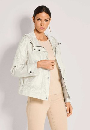 Frau mit dunklen Haaren in einer weißen Reißverschlussjacke über beigem Top und Hose, posierend vor hellem Hintergrund, trägt lange goldene Ohrringe.