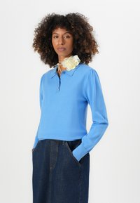 Lyseblå strikket pullover med krave og lange ærmer, parret med en denimnederdel; har en cremefarvet blomst som tilbehør ved halsen.