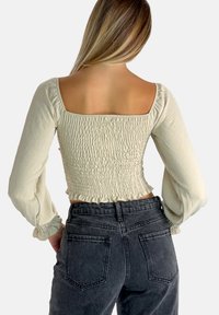 Ljust beige kort topp med ballongärmar, med en smockad design och volangkant. Matchad med högmidjade mörkgrå jeans.
