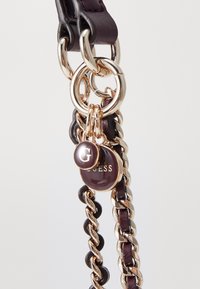 Læderrem med guldfarvet hardware, sammenflettede kæder og runde logotags med "G" og "Guess." Mørk farve med blanke accenter.