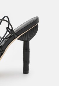 Sandale à talons hauts noire avec un talon en forme de coquillage texturé et de fines lanières tressées, dotée d'une semelle intérieure lisse et d'accents beiges.