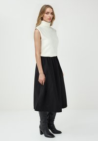 Donna che indossa un top a maniche corte bianco con collo alto, una gonna midi nera e stivali alti neri con tacco, in piedi contro uno sfondo bianco uniforme.