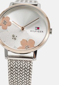 Montre en argent avec un cadran rond comportant trois accents floraux en rose et blanc, bracelet en mesh argenté et embellissements en cristal.
