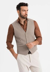 Mann mit dunklem Haar und Bart, der eine beigen Weste über einem braunen Hemd und weißen Hosen trägt und mit einer Hand in der Tasche posiert.