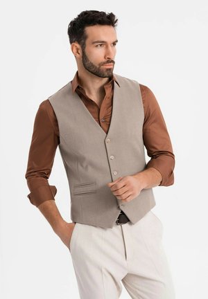 Mann mit dunklem Haar und Bart, der eine beigen Weste über einem braunen Hemd und weißen Hosen trägt und mit einer Hand in der Tasche posiert.