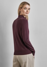 Pull-over rectangulaire bordeaux foncé, confectionné en tricot doux, avec un col montant et un ourlet côtelé, associé à un pantalon beige clair.