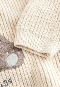 Tessuto a coste beige con un volto di animale ricamato in marrone e dettagli cuciti. La texture è morbida e pelosa.