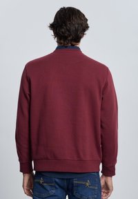 Sudadera de color burdeos con cuello redondo y mangas largas. Presenta puños y dobladillo acanalados. Lleva puesta sobre unos vaqueros con costuras amarillas en los bolsillos traseros.