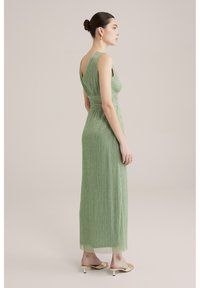 Pitkä vihreä mekko, jossa on pleated-muotoilu, tiukka yläosa ja avoin selkä. Läpinäkyvä päällinen ja nilkkapituinen helma lisäävät tyylikkyyttä.