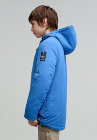 Chubasquero azul con capucha, de textura suave, bolsillo lateral y una etiqueta negra en la manga. El diseño general es simple y deportivo.