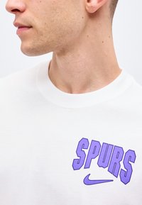 Jeune homme portant un t-shirt blanc avec le texte violet "SPURS" et un swoosh Nike violet sur la poitrine, présenté du cou vers le bas.