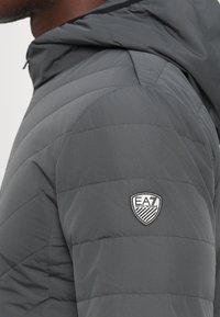 Detailný záber osoby v tmavošedej prešívanej bunde s logom EA7 Emporio Armani na hornej časti rukáva.