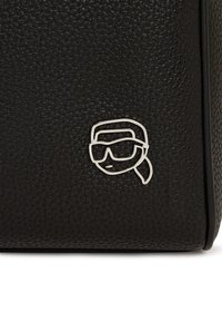 Sac en cuir noir avec une surface texturée et un logo de visage de cartoon argenté portant des lunettes de soleil. Le design présente une forme rectangulaire.