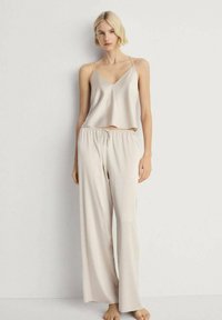 Bež satenasti pidžama set koji uključuje bezrukavni top s V-izrezom i široke hlače s elastičnim pojasom i vezicom.