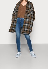 Chaqueta de cuadro en gris, negro y marrón, superpuesta a un suéter de punto marrón, combinada con jeans azules y zapatillas azul claro.