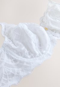 Soutien-gorge en dentelle blanche avec bords festonnés et motifs floraux complexes, présenté sur un fond neutre.