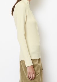 Langärmliges, hellgelbes, geripptes Oberteil mit einem halben Reißverschlusskragen und tailliertem Schnitt. Weicher Stoff, leicht dehnbares Material, minimalistische Details.