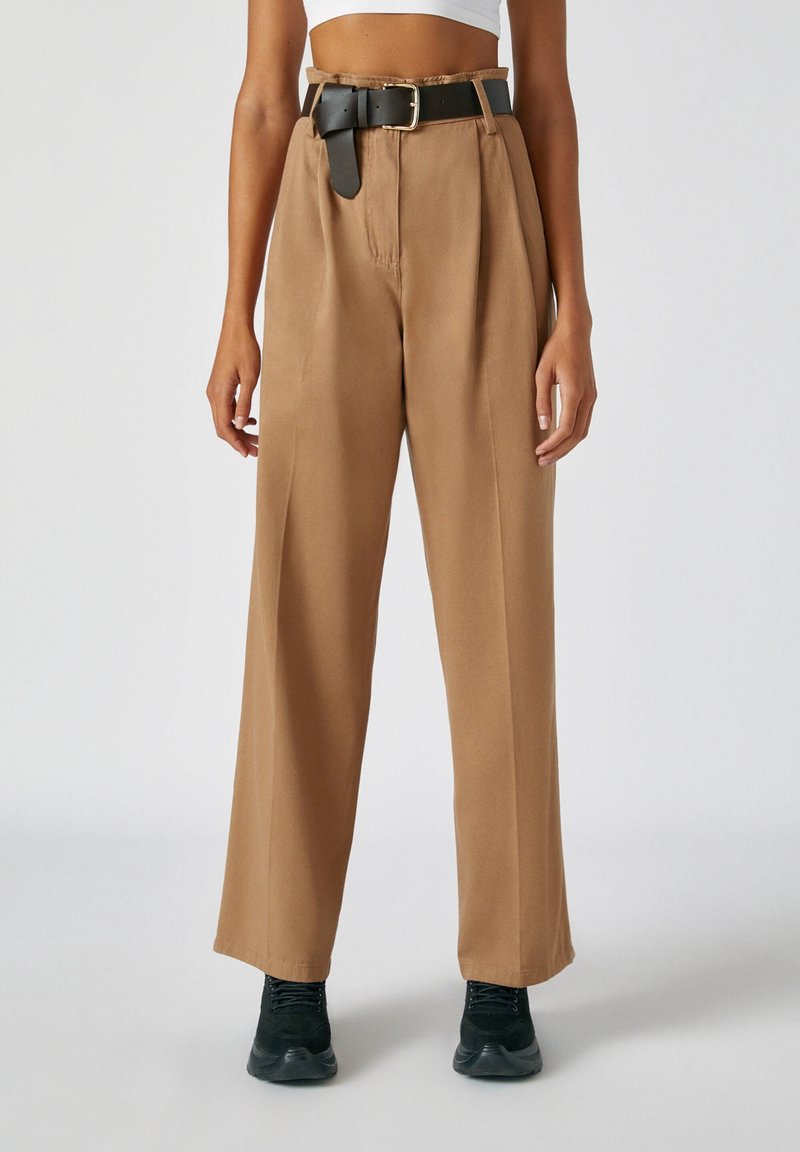 Femme portant un pantalon camel taille haute à jambes larges avec une ceinture noire et des baskets noires, un haut blanc court visible au-dessus de la taille.