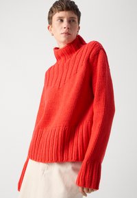 Roter, grob gestrickter Pullover mit hohem Kragen und gerippter Textur. Verfügt über breite Bündchen und eine lockere Passform, kombiniert mit hellen Hosen.