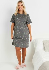 Camicia da notte grigia a maniche corte con motivi di stelle, lune e soli colorati. Il tessuto morbido arriva a metà coscia, abbinato a pantofole rosa soffici.