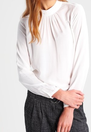 Biała bluzka z długim rękawem, z wysokim kołnierzykiem i marszczonym wykończeniem przy dekolcie, w połączeniu z ciemnymi, fakturowanymi spodniami.