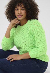 Gebreide trui in fel neon groen met een textuur in diamantenpatroon en geribde manchetten, gecombineerd met donkerblauwe spijkerbroek.