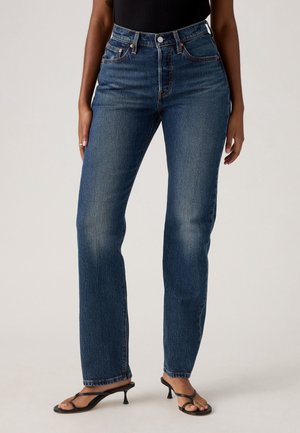 Person iført højtaljede, lige ben, mørkeblå jeans og sorte hælede sandaler, stående mod en ensfarvet baggrund.