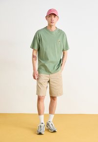 Mint green cotton t-shirt, beige shorts, pink cap, gray sneakers with white accents, standing on a yellow floor against a plain background.