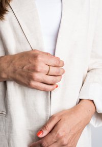 Blazer bej din in cu o cămașă albă dedesubt. Mâna are un inel subțire din aur, iar unghiile sunt vopsite în portocalie strălucitoare.