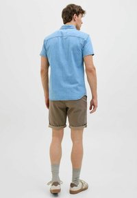 Lyseblå denim skjorte med korte ærmer, brune rullede shorts, grå strømper og hvide sneakers med brune detaljer. Ensfarvet baggrund.